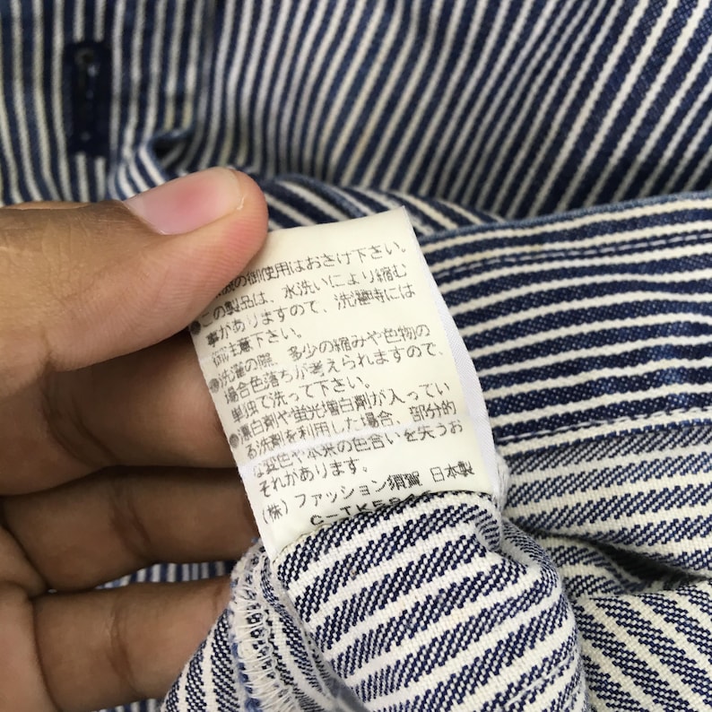 May include: Close-up of a blue and white striped fabric with a tag attached. The tag has Japanese text and care instructions. The fabric is a classic striped pattern, likely for clothing.