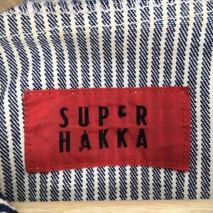 May include: Close-up of a red tag with the text "SUPER HAKKA" in black letters, sewn onto a blue and white striped fabric. The fabric has a textured appearance.