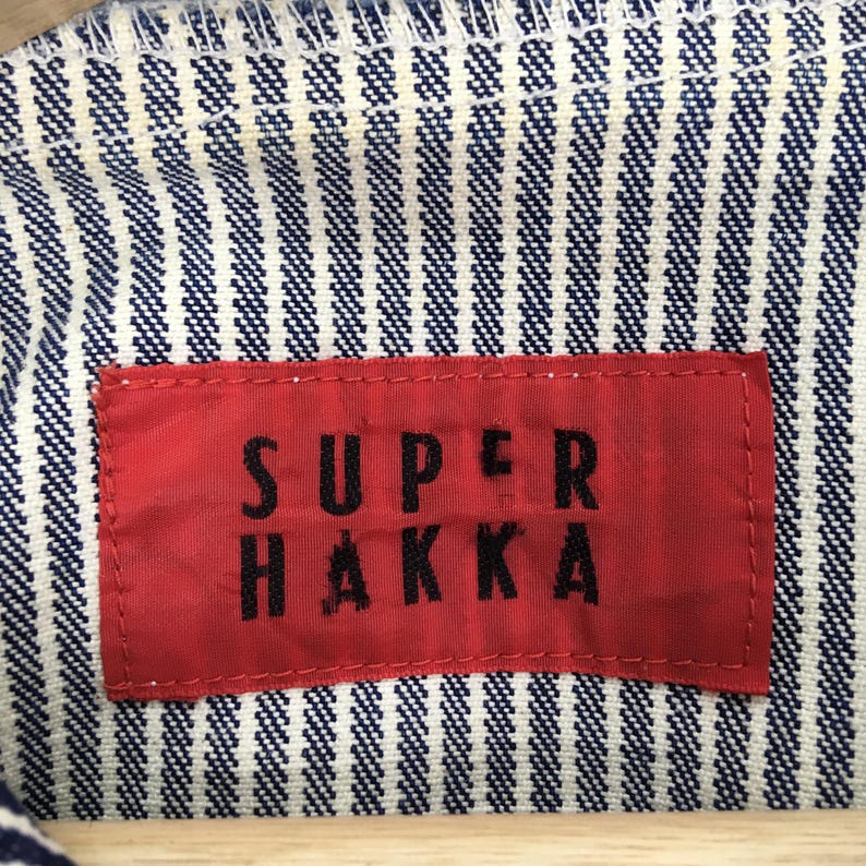 May include: Close-up of a red tag with the text "SUPER HAKKA" in black letters, sewn onto a blue and white striped fabric. The fabric has a textured appearance.