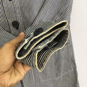 May include: Close-up of a blue and white striped jacket with black buttons. The sleeve is folded up, revealing the interior lining and the cuff. The jacket has a pocket and is made of a striped fabric.
