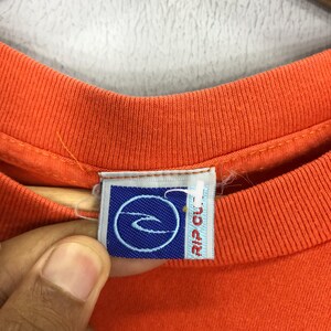 Pu&ograve; includere: T-shirt arancione con un'etichetta logo blu e bianca che dice "Rip Curl".