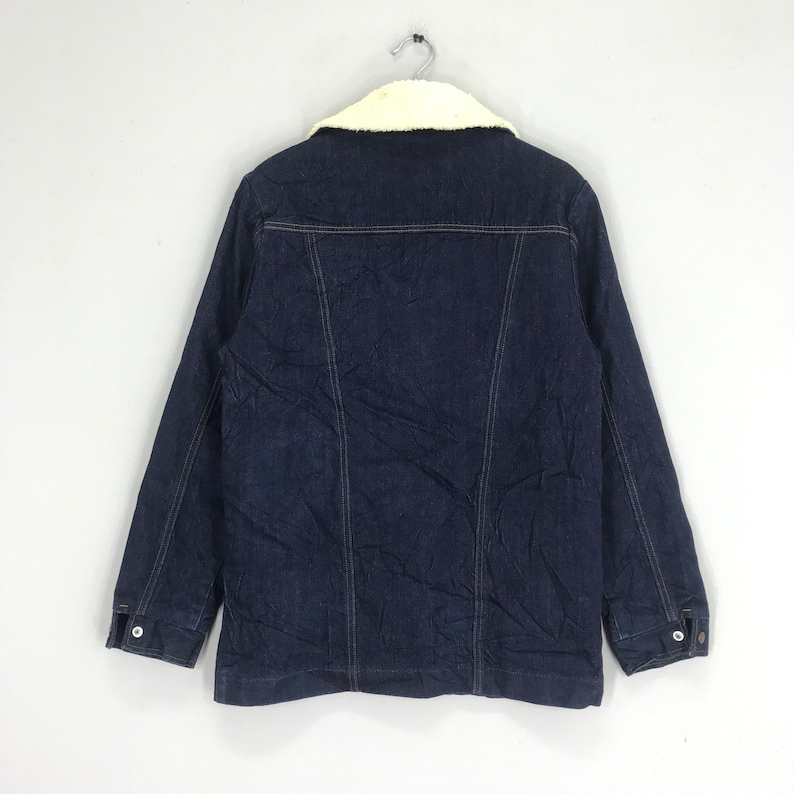 Op de afbeelding: Een donkerblauw denim jack met een witte nep bontkraag. Het jack heeft een knoopsluiting en twee zakken aan de voorkant.