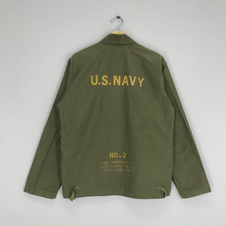 May include: Olive green jacket with the text "U.S. NAVY" printed in gold on the back. The jacket also has additional text and numbers printed below the main text. The jacket is hanging on a metal hanger.