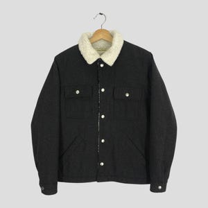 Puede incluir: Chaqueta gris oscuro con cuello y forro de color crema y peludo. La chaqueta tiene dos bolsillos en el pecho, dos bolsillos laterales y botones plateados. La chaqueta está colgada de una percha de madera.