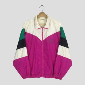 Op de afbeelding: Een vintage windjack in wit, fuchsia, zwart en groen. De jas heeft een volledige ritssluiting, een kraag en een logo op de borst. Het colorblock-ontwerp heeft een chevronpatroon over de borst en mouwen. De jas hangt aan een houten hanger.