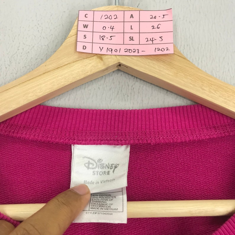 Puede incluir: Sudadera rosa con una etiqueta blanca de Disney Store. La etiqueta tiene el texto "Disney Store" y "Hecho en Vietnam". La etiqueta tambi&eacute;n indica la composici&oacute;n de la tela como 80% algod&oacute;n y 20% poli&eacute;ster. El n&uacute;mero de estilo es 571042044.