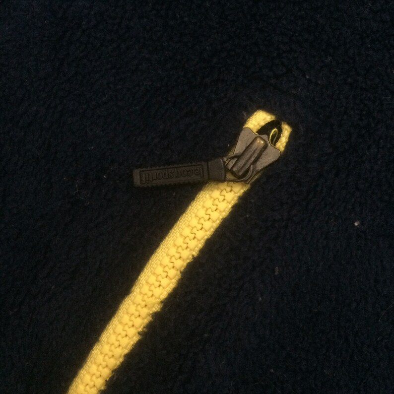 Op de afbeelding: Close-up van een gele rits met een zwarte rits-trekker waarop "Jack Sport" staat. De rits zit op een donkerblauwe stof.