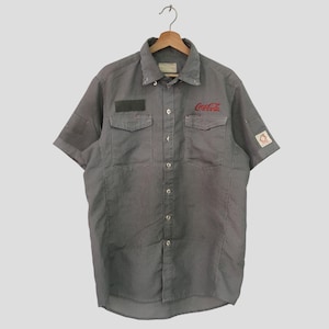 May include: A gray and white striped short-sleeved button-down shirt with a red Coca-Cola logo embroidered on the front. The shirt has two chest pockets and a small patch on the sleeve.