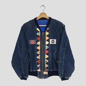 Puede incluir: Chaqueta bomber de mezclilla azul con forro azul y cuello y puños azul marino. La chaqueta presenta un colorido diseño de patchwork en el centro y dos parches en la parte delantera. Una cremallera dorada recorre la parte delantera.