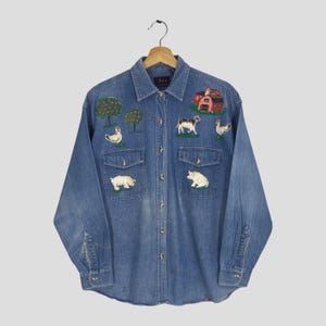 May include: Blue denim shirt with farm animal embroidery. The design includes a red barn, trees, and farm animals such as a cow, pig, and chicken. Long-sleeved with two chest pockets.