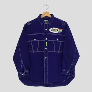 Puede incluir: Una camisa de manga larga azul oscuro con botones, costuras blancas en contraste, dos bolsillos en el pecho y un parche de Benetton Formula 1 en el lado derecho. La prenda incluye una pequeña etiqueta verde en la tapeta central y charreteras.