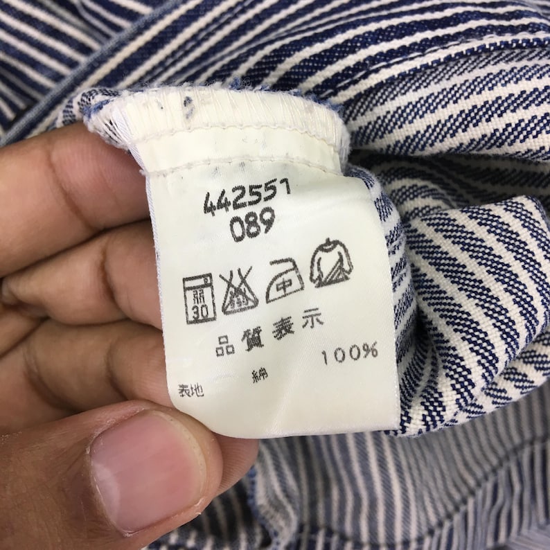 May include: Close-up of a white tag with black text and symbols, attached to a blue and white striped fabric. The tag includes numbers, care symbols, and the text "100%" in black.