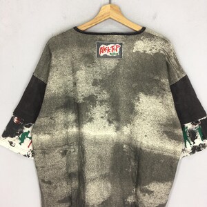 May include: An oversized vintage t-shirt with a distinctive, distressed design. The shirt has a faded, mottled pattern in shades of grey and black. The sleeves are a darker grey, and the cuffs have a colourful graphic. The "Black Top" Reebok patch is visible.