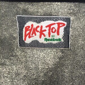 May include: Close-up of grey fabric with a rectangular patch. The patch has a black mesh border and features the words "BLACK TOP" in red, with "by Reebok" in green.