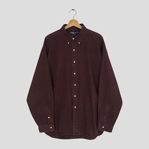 May include: A long-sleeved, button-down shirt in a deep burgundy color. The shirt features a button-down collar, a small embroidered logo on the chest, and buttoned cuffs. It is hanging on a wooden hanger against a light gray background.