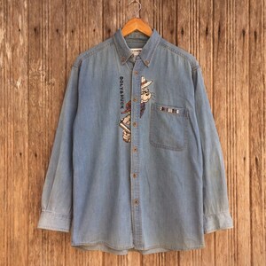 Puede incluir: Camisa vaquera azul claro con botones y mangas largas. Presenta un gráfico de perro de dibujos animados con el texto "Doly & Huck" y "Doly & G" en el bolsillo. La camisa está colgada en una percha de madera.