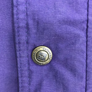 May include: Close-up of a purple fabric with a button. The button is circular, metallic, and features the brand name "Columbia" and a logo. The fabric has visible stitching and a textured appearance.