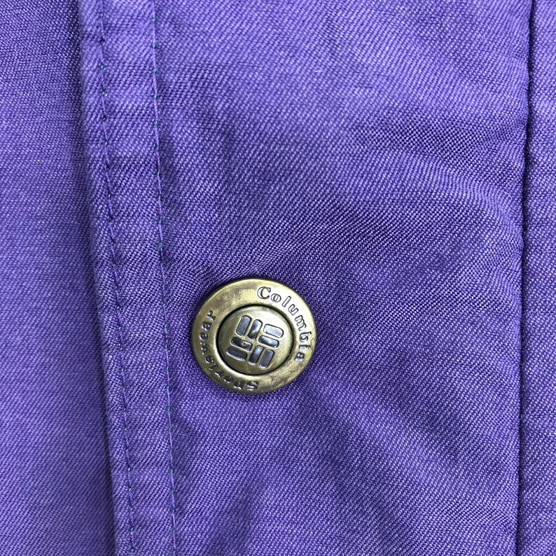 May include: Close-up of a purple fabric with a button. The button is circular, metallic, and features the brand name "Columbia" and a logo. The fabric has visible stitching and a textured appearance.