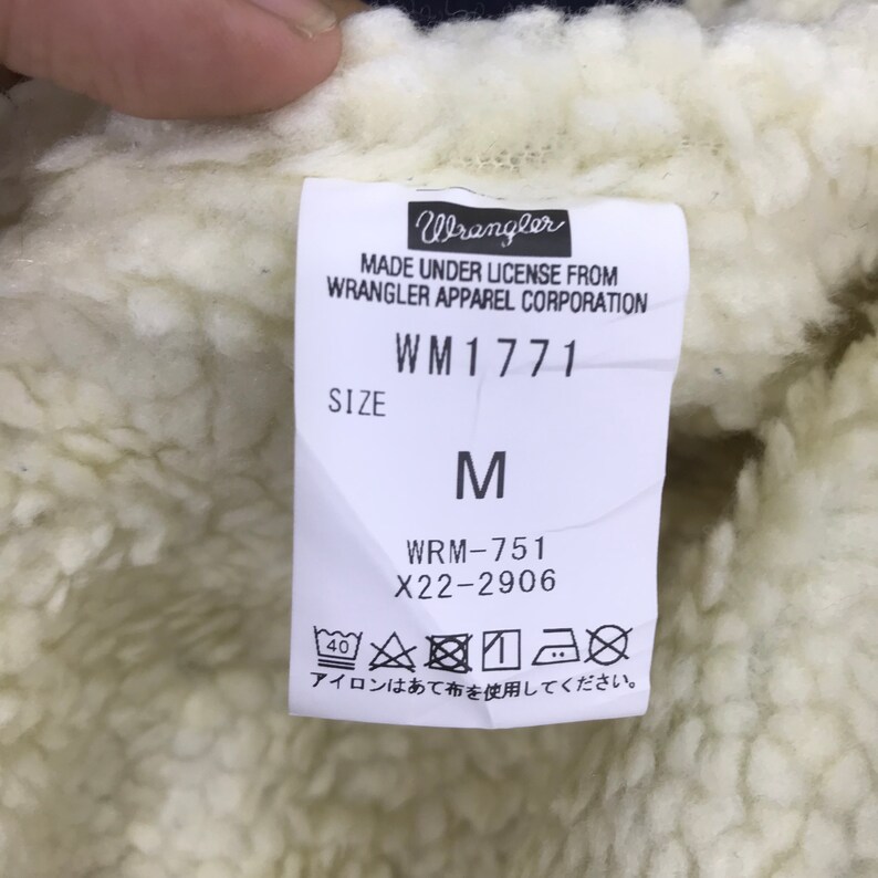 Op de afbeelding: Een wit label met zwarte tekst dat "Wrangler" en "Made under license from Wrangler Apparel Corporation" zegt. Het label bevat ook het modelnummer "WM1771", maat "M" en stylenummer "WRM-751 X22-2906". Het label heeft een witte achtergrond en zwarte tekst.