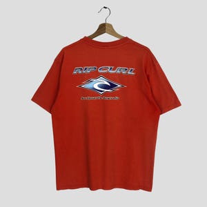 Pu&ograve; includere: T-shirt rosso a maniche corte con un logo Rip Curl blu e bianco sul retro. Il logo presenta un'onda e il testo "Surfboards Australia".