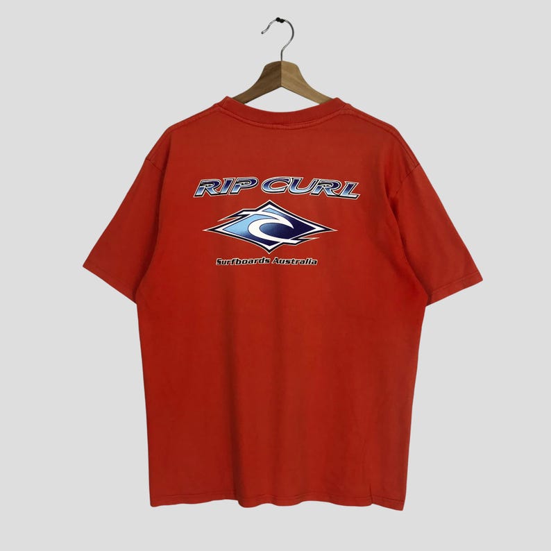 Pu&ograve; includere: T-shirt rosso a maniche corte con un logo Rip Curl blu e bianco sul retro. Il logo presenta un'onda e il testo "Surfboards Australia".