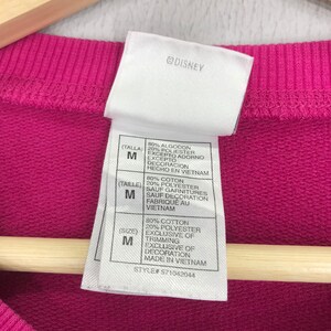Puede incluir: Una sudadera rosa de mezcla de algod&oacute;n y poli&eacute;ster con una etiqueta blanca. La etiqueta tiene el texto "@DISNEY" y detalla la composici&oacute;n del tejido, la talla y el origen. La etiqueta tambi&eacute;n incluye el texto "(Talla) M", "(Taille) M" y "(Size) M".