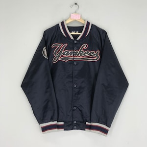 Può includere: Una giacca da baseball in satin blu navy con un logo "Yankees" ricamato in rosso e bianco sul davanti. La giacca ha un colletto, polsini e orlo a coste. Ha una chiusura con bottoni e due tasche.