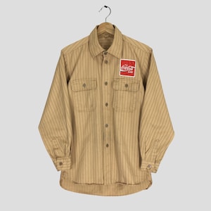 May include: A tan and brown striped long-sleeved shirt with a button-up front and two chest pockets. The shirt has a Coca-Cola logo patch on the right chest pocket.