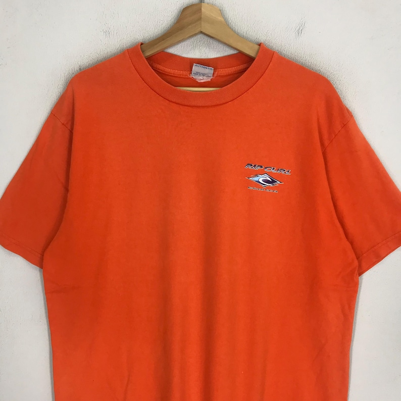 Pu&ograve; includere: Maglietta arancione a maniche corte con un logo Rip Curl che presenta un'onda e il testo "Rip Curl" e "Australia's Own".