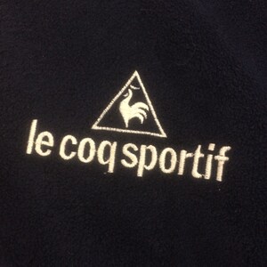 Op de afbeelding: Een marineblauwe fleecejas met een wit geborduurd logo van een haan in een driehoek en de tekst "le coq sportif".