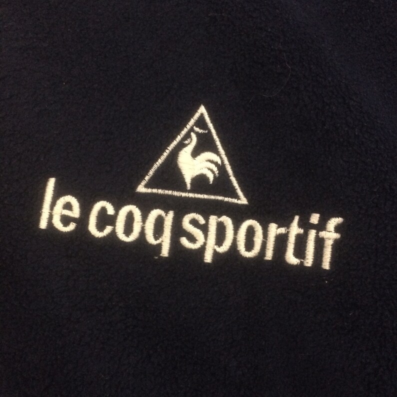 Op de afbeelding: Een marineblauwe fleecejas met een wit geborduurd logo van een haan in een driehoek en de tekst "le coq sportif".