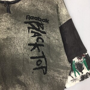 May include: A vintage Reebok Black Top t-shirt in grey with a speckled pattern. The shirt displays the Reebok logo and "BLACK TOP" in large black lettering. The sleeve has a black mesh panel and a painted design.