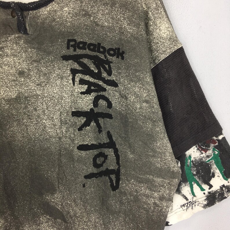 May include: A vintage Reebok Black Top t-shirt in grey with a speckled pattern. The shirt displays the Reebok logo and "BLACK TOP" in large black lettering. The sleeve has a black mesh panel and a painted design.