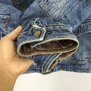 Puede incluir: Primer plano de una chaqueta vaquera azul con forro de pana marr&oacute;n. La chaqueta tiene cierre de bot&oacute;n y un solo bolsillo en el pecho.