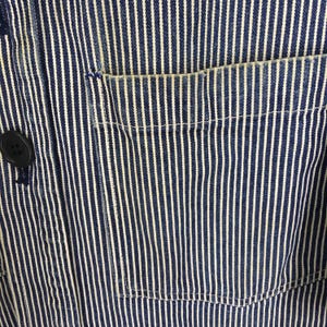May include: Close-up of a blue and white striped fabric with a pocket and a black button. The fabric has vertical white stripes against a blue background. The pocket is on the right side of the image.