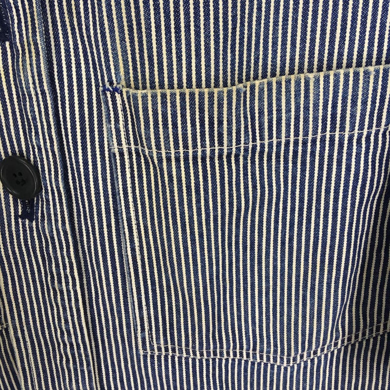 May include: Close-up of a blue and white striped fabric with a pocket and a black button. The fabric has vertical white stripes against a blue background. The pocket is on the right side of the image.