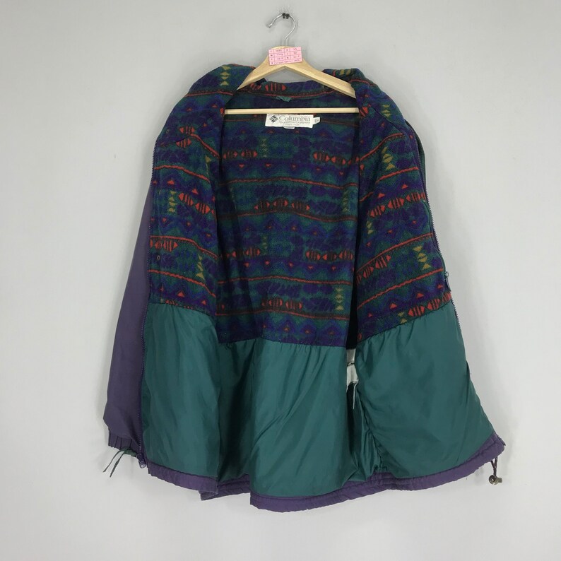 May include: A vintage Columbia jacket, featuring a purple exterior and a green lower section. The interior showcases a patterned fleece lining in shades of green, blue, and orange. The jacket is hanging on a wooden hanger.