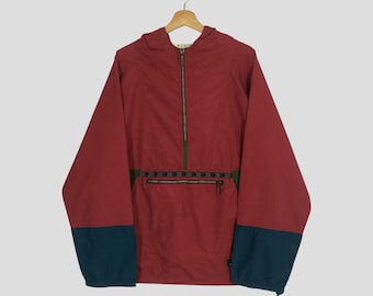 Peut inclure: Veste anorak rouge avec capuche et fermeture éclair partielle. Les poignets sont bleu sarcelle, et une bande à motifs orne la poitrine. La veste est suspendue à un cintre en bois.