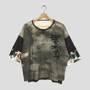 May include: Vintage Reebok Black Top t-shirt with a distinctive, distressed pattern. This crew neck t-shirt has short sleeves and the Reebok logo with "Black Top" printed vertically. The sleeves have a black panel and a colourful graphic.