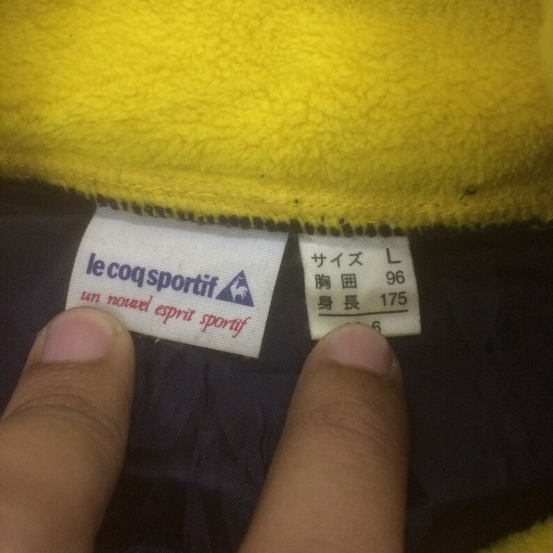 Op de afbeelding: Een blauw en geel kledinglabel met de tekst "le coq sportif" en "un nouvel esprit sportif". Het label heeft ook een haanlogo. Een wit label met Japanse tekst en maat L, borstomvang van 96 cm en lengte van 175 cm.