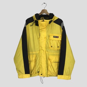 Puede incluir: Chaqueta Polo Sport amarilla y negra con capucha. La chaqueta tiene rayas negras en los laterales y un pequeño bolsillo con un logotipo. Cuelga de una percha de madera.