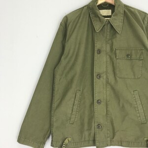 May include: Olive green jacket with a button-up front, a collared neckline, and a single chest pocket. The jacket has two lower pockets and a strap detail at the hem. The jacket is made of a durable fabric.