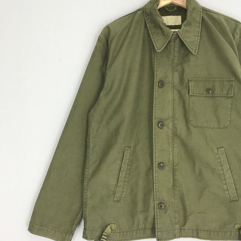 May include: Olive green jacket with a button-up front, a collared neckline, and a single chest pocket. The jacket has two lower pockets and a strap detail at the hem. The jacket is made of a durable fabric.