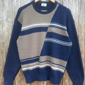 May include: A blue and brown striped sweater with a green crocodile logo. The sweater has a crew neck and long sleeves.