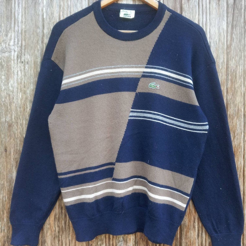May include: A blue and brown striped sweater with a green crocodile logo. The sweater has a crew neck and long sleeves.