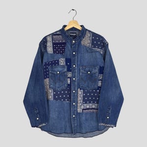 Könnte beinhalten: Ein langärmeliges Jeanshemd von Wrangler mit einem Patchwork-Design aus dunkelblauen und weißen Bandana-Mustern. Das Hemd verfügt über eine durchgehende Knopfleiste, einen Kragen und zwei Brusttaschen mit Druckknöpfen auf einem mittelblauen Stoff.