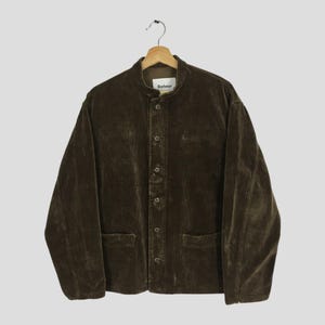 May include: A brown corduroy jacket with a button-up front, two front pockets, and a mandarin collar. The jacket is hanging on a wooden hanger. The brand name "Barbour" is visible on the collar.