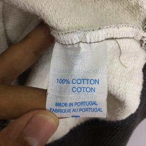 May include: A close-up of a white fabric label with blue text. The label states "100% COTTON COTON MADE IN PORTUGAL FABRIQUE AU PORTUGAL". The label is attached to a light grey and white garment.