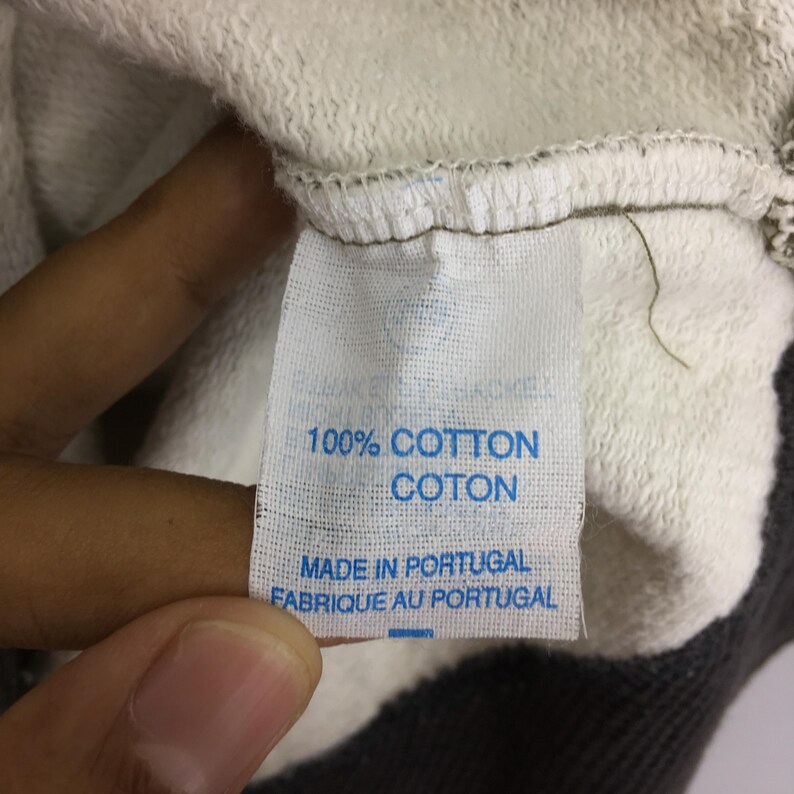 May include: A close-up of a white fabric label with blue text. The label states "100% COTTON COTON MADE IN PORTUGAL FABRIQUE AU PORTUGAL". The label is attached to a light grey and white garment.