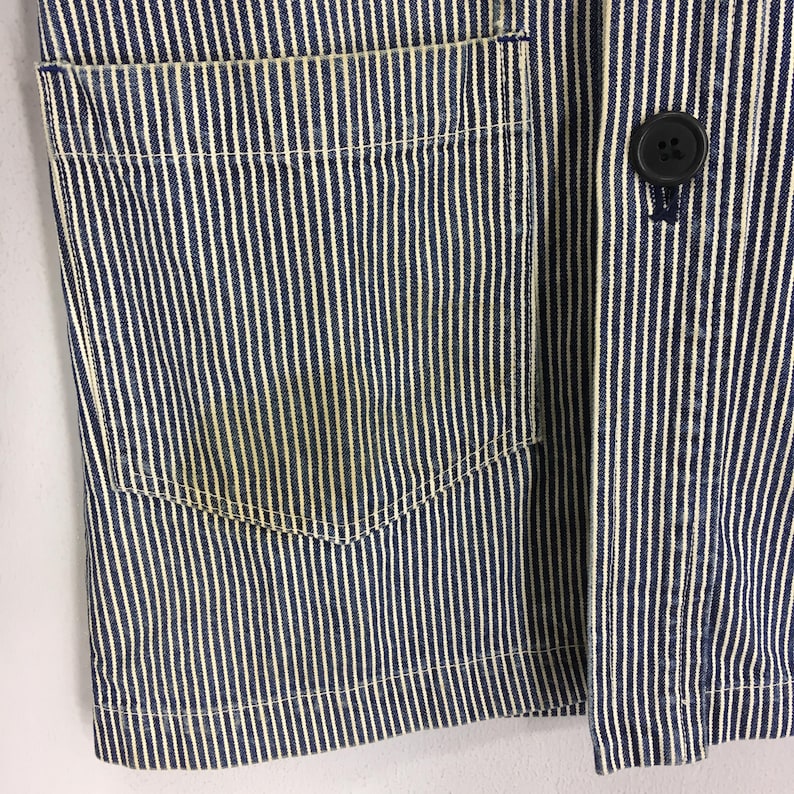 May include: Close-up of a blue and white striped jacket. The jacket features a pocket on the left side and a black button closure. The fabric has a vertical stripe pattern.